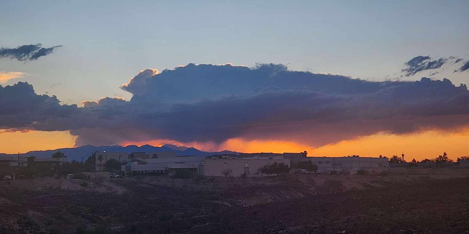 A dark storm cloud dropping rain over desert mountains at sunset, above a bare dirt lot and a row of low buildings